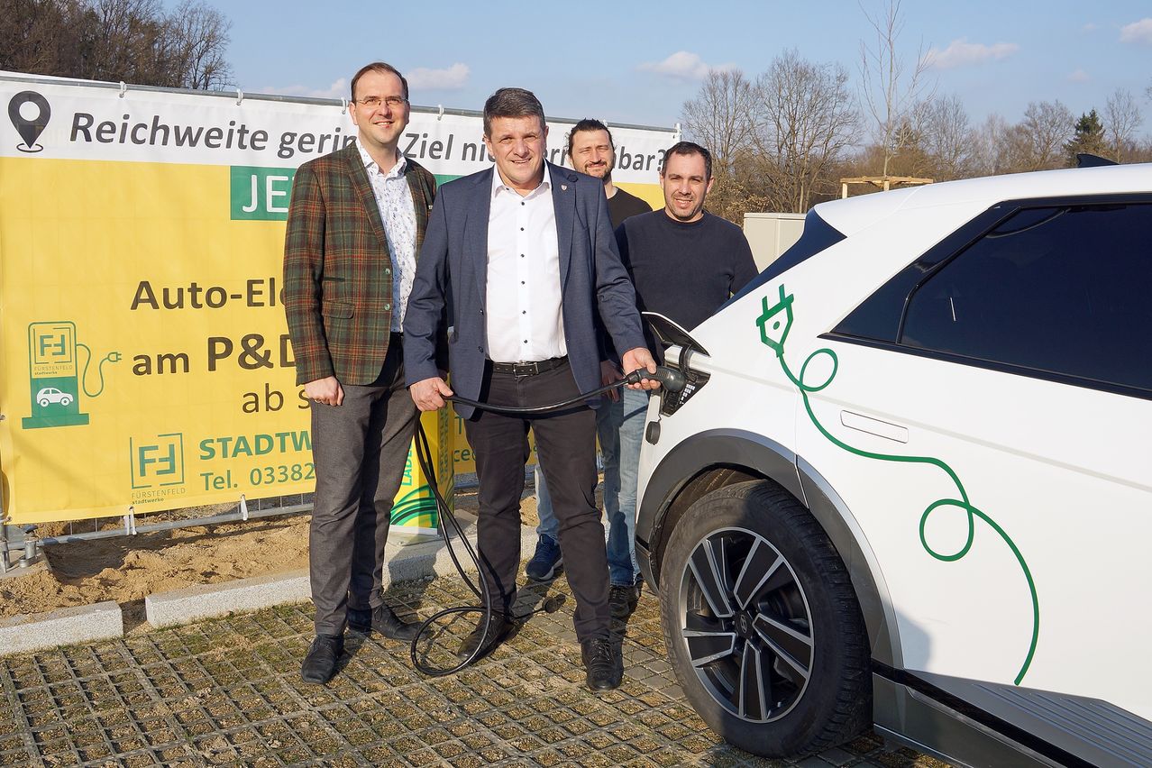 Four men stand by a white electric car with a charging cable plugged in, on a brick pavement. Behind them is a yellow banner.