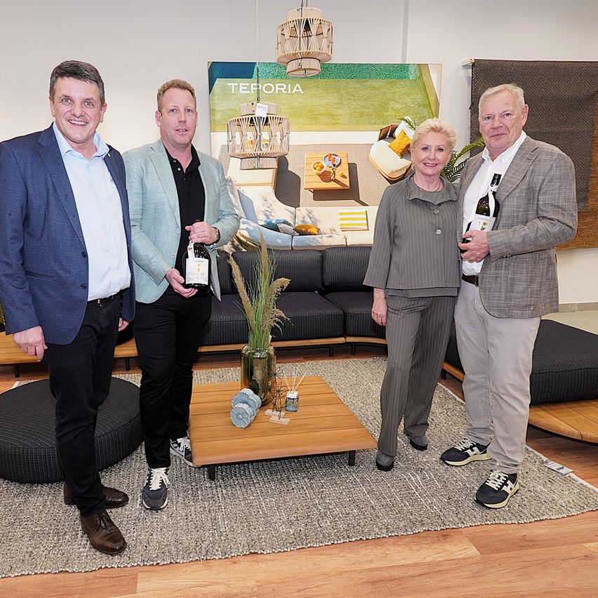 Four adults stand in a living room holding wine bottles, smiling. They are in front of a couch and a table with a vase and other items.