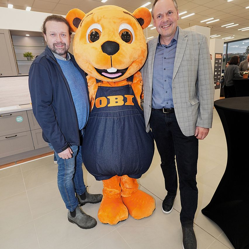 Two men stand in an indoor space, one wearing a blue jacket and jeans, the other in a gray suit. They are both smiling and posing with a large orange bear mascot that has the word Obi written on its apron.