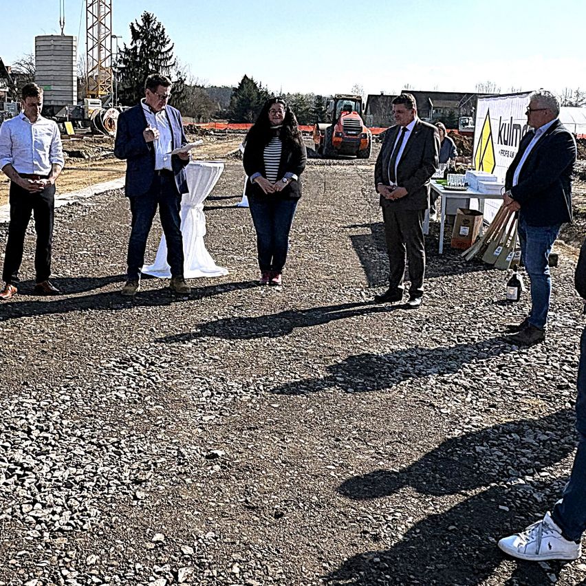 Six people stand on a gravel surface. A man reads from a paper. A woman stands with her hands clasped in front of her. Equipment and a house are in the background.