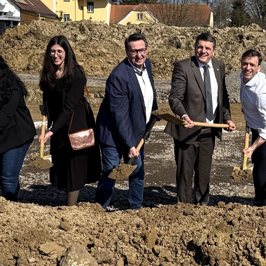 Five individuals, four men and one woman, are at a construction site, smiling and holding shovels, perhaps for a groundbreaking ceremony. Behind them are piles of dirt and a yellow house.