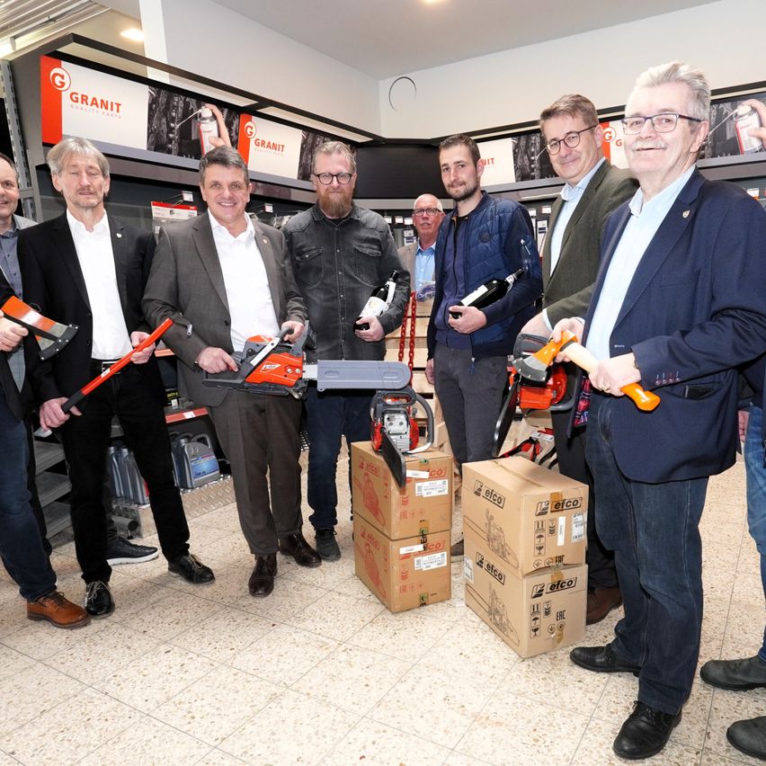 Eine Gruppe von Männern in Anzügen und mit Brillen posiert für ein Foto in einem Geschäft mit Werkzeugen und Kisten auf dem Boden.