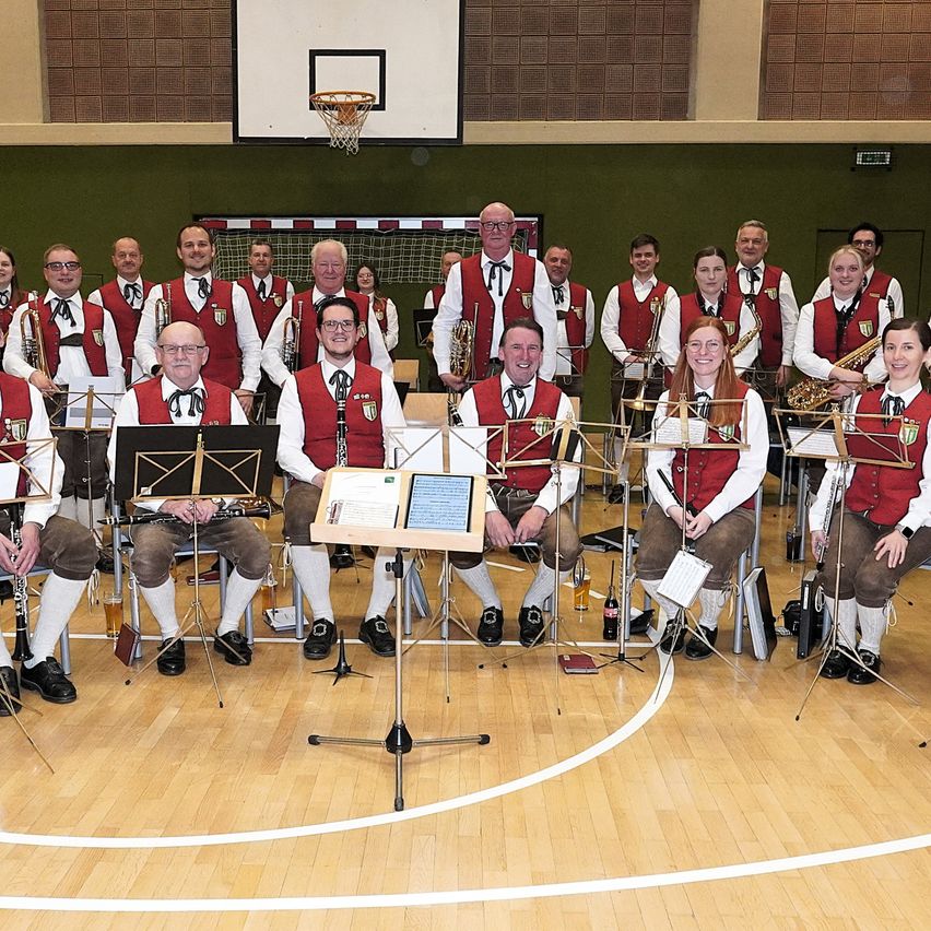 Eine Gruppe von Musikern in traditioneller Kleidung posiert für ein Foto in einer Sporthalle. Sie sitzen mit verschiedenen Musikinstrumenten. Die Sporthalle hat einen Basketballkorb im Hintergrund.