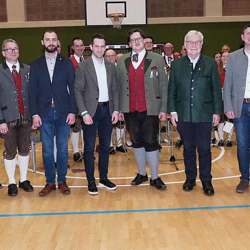 Gruppe von Männern in traditioneller deutscher Kleidung, die in einem Gymnasium stehen, möglicherweise für eine Preisverleihung. Sie sind in verschiedenen Stilen von Anzügen und traditioneller bayerischer Kleidung gekleidet.