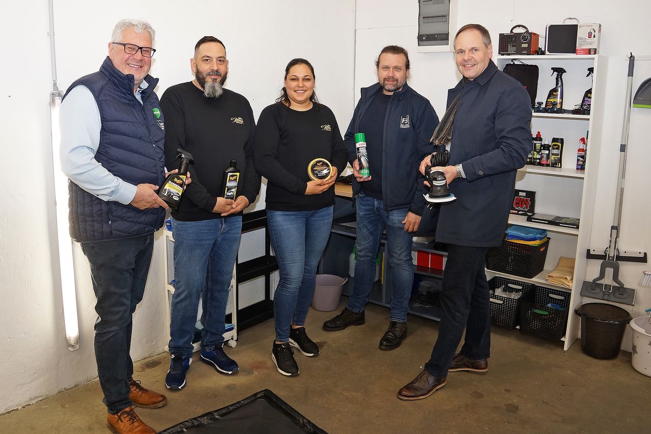 Fünf Personen stehen in einer Garage, halten Produkte und lächeln. Sie sind von Regalen mit Gegenständen und einem Mülleimer umgeben.