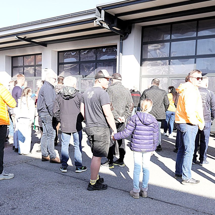Eine Gruppe von Menschen steht vor einem Gebäude mit großen Glasfenstern. Einige tragen Hüte und Brillen. Ein Mädchen in einer lila Jacke hält die Hand eines Mannes.