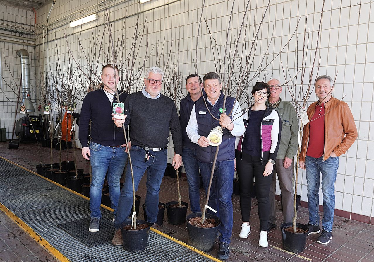 Eine Gruppe von Menschen steht in einer Baumschule und hält kleine Topfpflanzen. Sie lächeln und scheinen für ein Foto zu posieren. Einige tragen Brillen und Jacken.