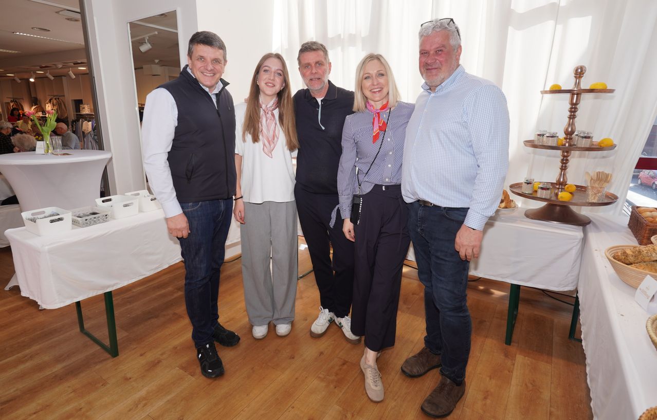 Fünf Personen stehen zusammen in einem Raum und lächeln für ein Foto. Zwei Frauen und drei Männer, lässig gekleidet, scheinen für ein Gruppenfoto zu posieren. Der Raum hat weiße Wände, Holzböden und weiße Vorhänge.