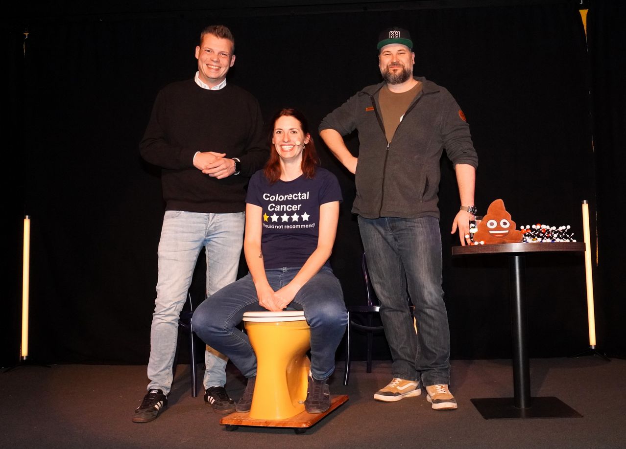 Drei Personen stehen auf einer Bühne, zwei Männer und eine Frau. Die Frau sitzt auf einem gelben Trommel und lächelt. Der Mann rechts trägt eine Kappe und eine Uhr.