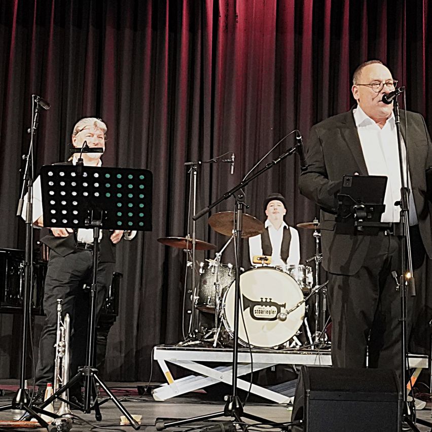 Eine Band tritt auf der Bühne auf. Der Leadsänger steht vor einem Mikrofon, mit einem Schlagzeuger und einem Trompeter im Hintergrund. Dahinter ist ein roter Vorhang als Hintergrund.