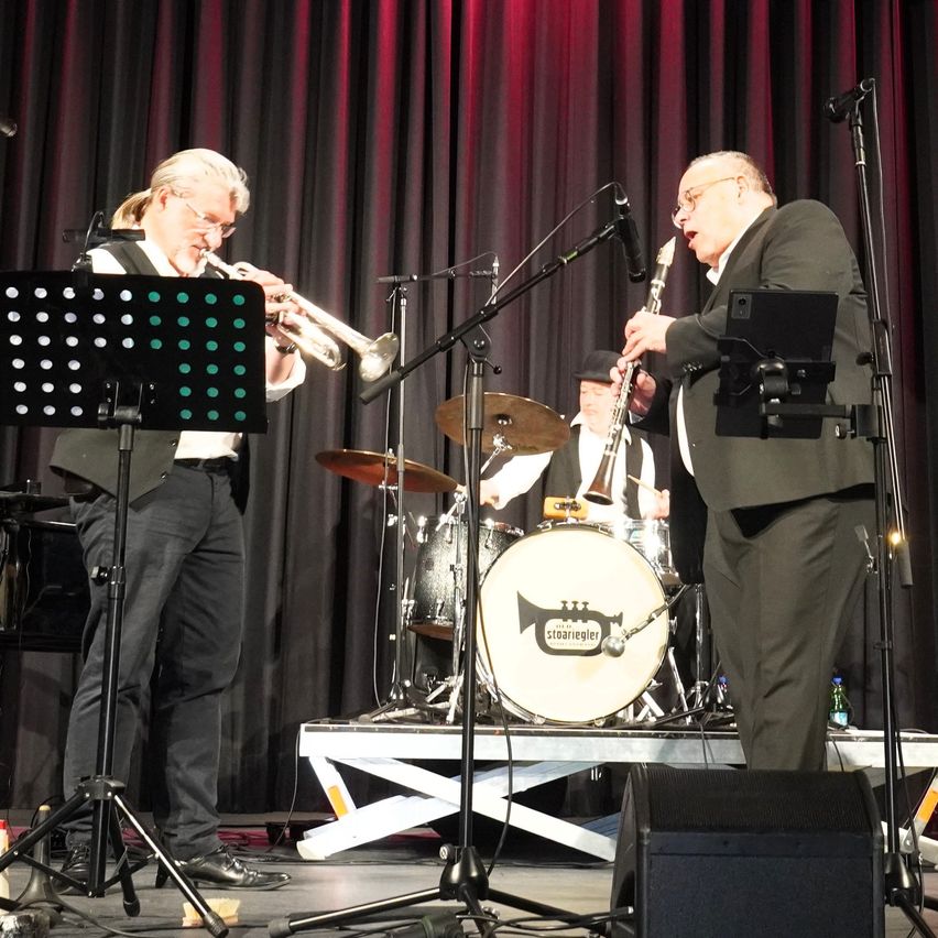 Drei Musiker spielen auf der Bühne, einer spielt Trompete, ein anderer Klarinette und ein Dritter Schlagzeug. Sie stehen vor Mikrofonen, mit einem Schlagzeug und Becken dahinter.