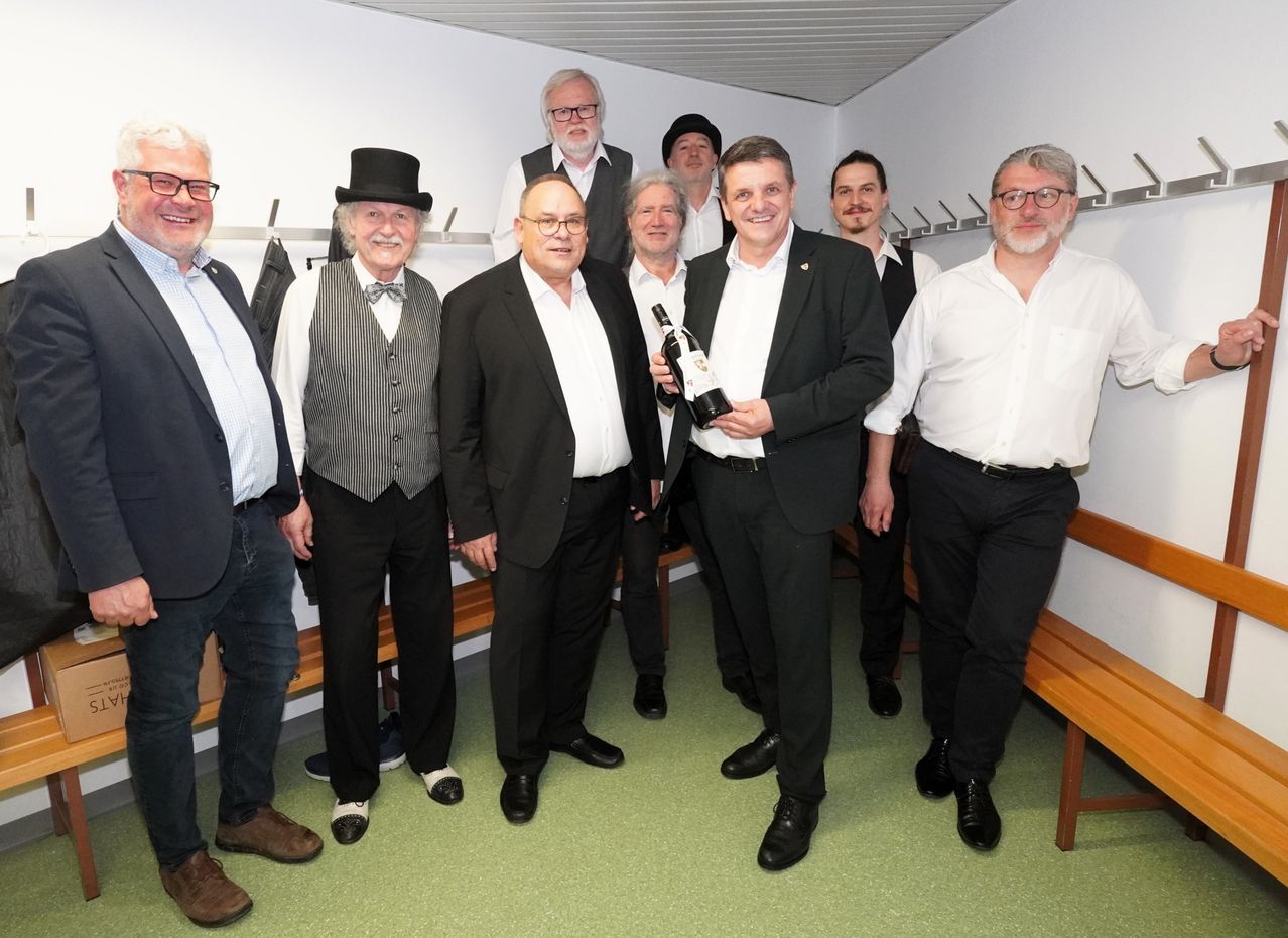 Eine Gruppe von Männern in formeller Kleidung posiert für ein Foto in einer Umkleidekabine mit einem Mann, der eine Flasche Wein hält.