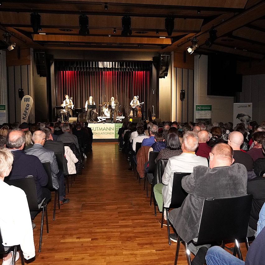 Eine Band spielt auf der Bühne in einem Saal mit Holzboden, rotem Vorhang als Hintergrund und einem Publikum, das in Reihen sitzt. Die Bühne hat Lautsprecher, Mikrofone und Instrumente. Es gibt Banner mit Logos und Text an den Seiten der Bühne.
