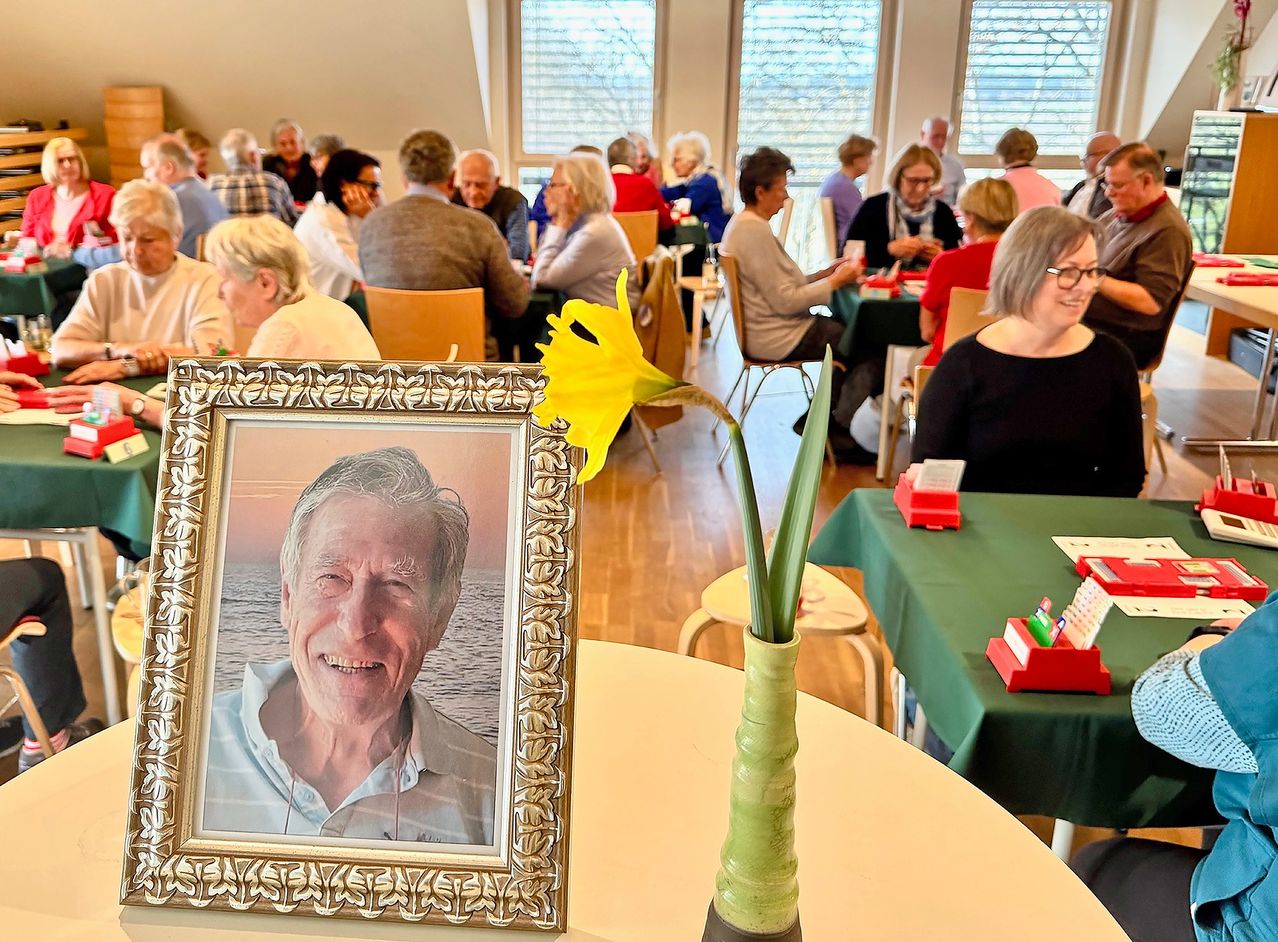 Ein Foto eines lächelnden älteren Mannes steht auf einem Tisch in einem Essbereich. Ein Vase mit gelben Blumen steht neben dem Foto. Viele Menschen sitzen an Tischen, einige unterhalten sich.