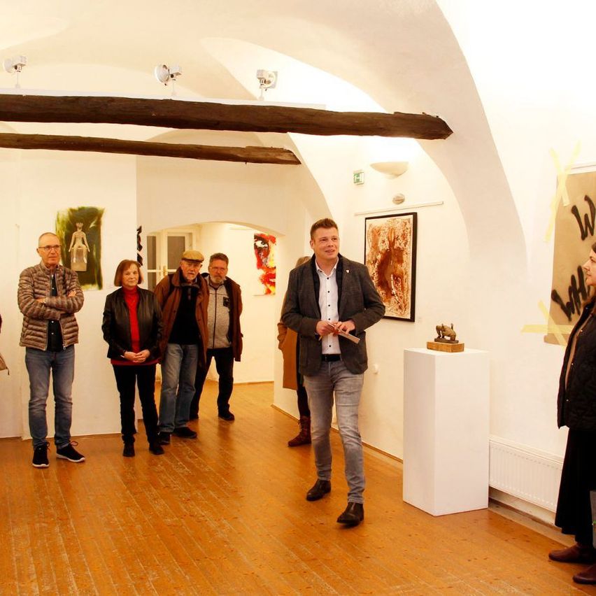 Eine Gruppe von Menschen steht in einer Kunstgalerie, wobei ein Mann spricht und andere zuhören. Der Raum hat Holzböden, gewölbte Decken und Wände, die mit Gemälden und Skulpturen geschmückt sind.