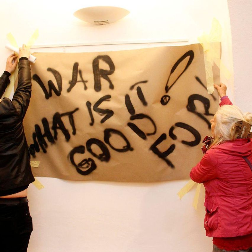 Zwei Personen hängen ein braunes Banner an einer Wand auf. Es hat schwarze Graffiti mit den Worten 'WAR WAS IST GUT DAFÜR?' darauf geschrieben.