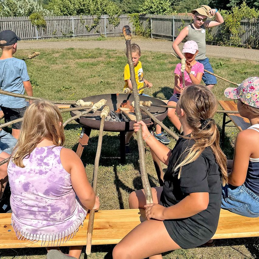 Eine Gruppe von Kindern und Erwachsenen versammelt sich um ein Feuerloch und röstet Marshmallows auf Stöcken. Einige sitzen auf Holzbänken, während andere in der Nähe stehen. Der Ort ist im Freien, mit einem Holzzaun und Grünpflanzen im Hintergrund.