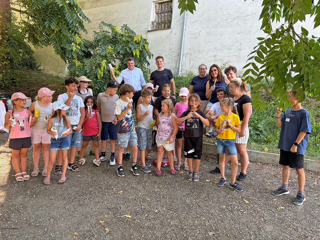 Eine Gruppe von Menschen, darunter Kinder und Erwachsene, steht vor einem Gebäude mit viel Grün. Sie halten Eis und posieren für ein Foto.