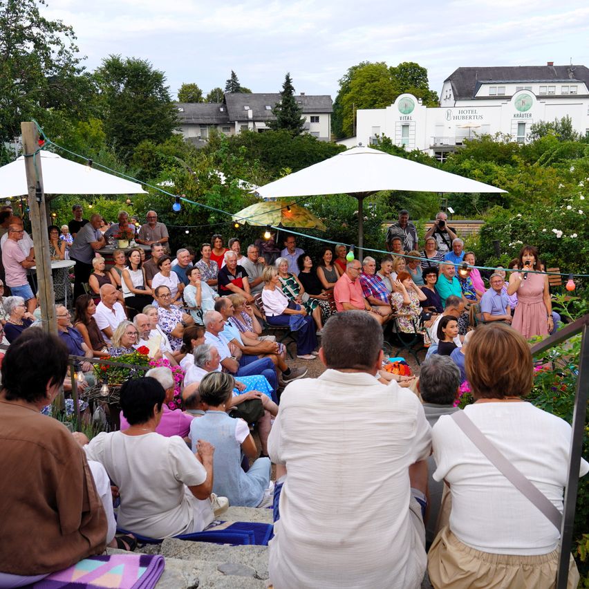 Eine Gruppe von Menschen versammelt sich in einem Garten, einige sitzen auf dem Boden und andere stehen, mit einer Frau, die unter einem Schirm in ein Mikrofon spricht. Bäume und Gebäude sind im Hintergrund.