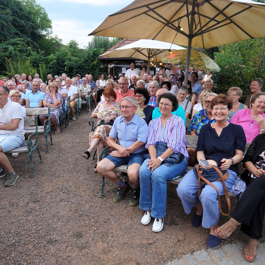 Eine große Gruppe von Menschen sitzt in Reihen unter Sonnenschirmen in einer Gartenumgebung im Freien. Sie sind lässig gekleidet und scheinen eine gesellige Zusammenkunft zu genießen.