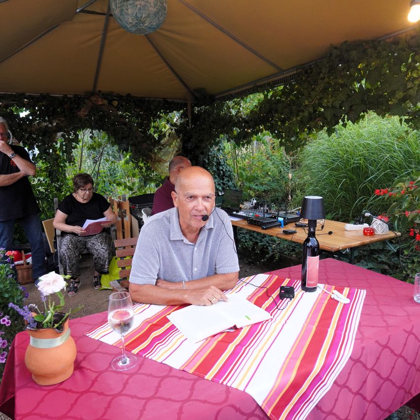 Ein Mann sitzt mit einem Mikrofon an einem Tisch, umgeben von einer Gartenumgebung. Eine Weinflasche und ein Glas stehen auf dem Tisch.