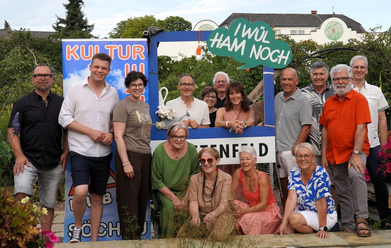 Eine Gruppe von Menschen posiert für ein Foto vor einem Schild mit der Aufschrift 'I wül HAM NOCH' und einem weiteren Schild mit dem Wort 'Kultur'. Sie alle lächeln und scheinen sich zu amusieren.