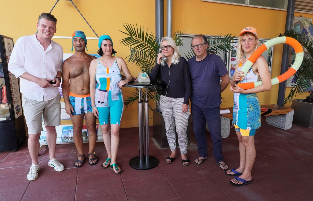 Eine Gruppe von Menschen in bunter Strandkleidung steht in einem Raum, eine Frau telefoniert.