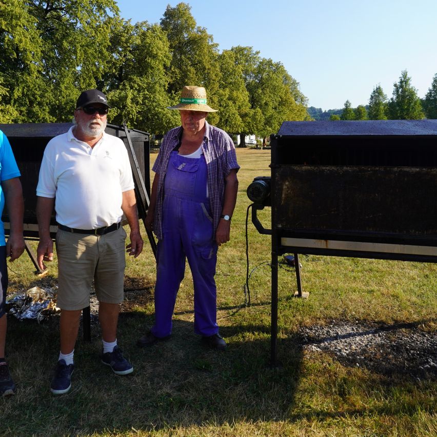 Zwei Männer in Freizeitkleidung stehen neben einem Grill auf einer Wiese mit Bäumen im Hintergrund.