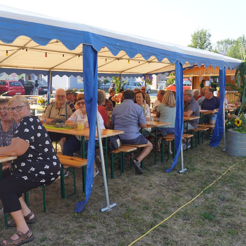 Eine Gruppe von Menschen sitzt unter einem blauen Zelt an Tischen und genießt Essen und Getränke. Das Zelt ist mit blauen Vorhängen dekoriert.