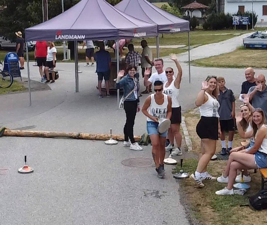 Eine Gruppe von Menschen versammelt sich unter einem violetten Zelt mit dem Wort 'LANDMANN' darauf. Einige stehen und winken, während andere auf dem Gras sitzen. Eine Frau in der Mitte hält eine Frisbee.