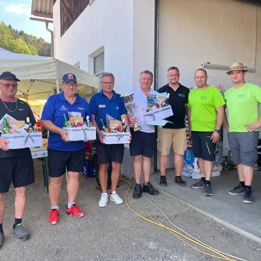 Fünf Männer stehen vor einem Gebäude und halten Trophäen und Geschenkkörbe. Sie sind in Freizeitkleidung gekleidet, und einer der Männer trägt einen Hut. Hinter ihnen ist ein Zelt aufgestellt, und ein Banner ist sichtbar.
