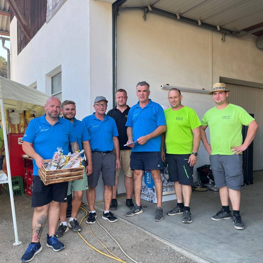 Eine Gruppe von Männern in passenden blauen und grünen Shirts steht vor einem Gebäude, einer hält einen Korb mit eingewickelten Gegenständen.