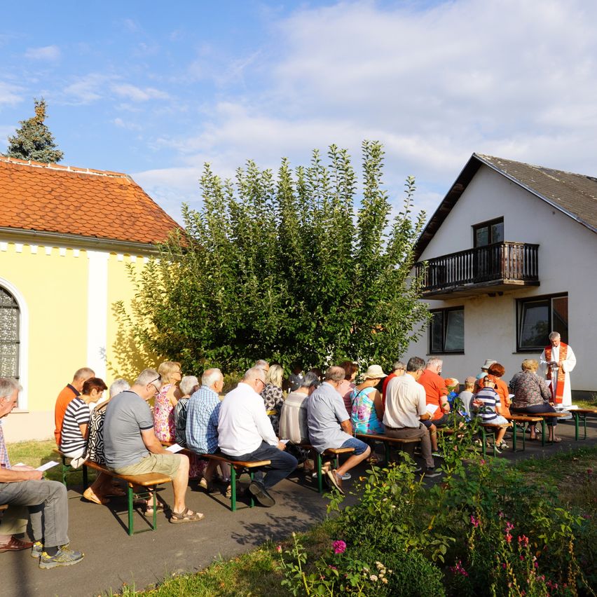 Eine Gruppe von Menschen sitzt auf Bänken in einem Garten und hört einem Priester zu. Der Hintergrund zeigt ein Haus und einen Baum.