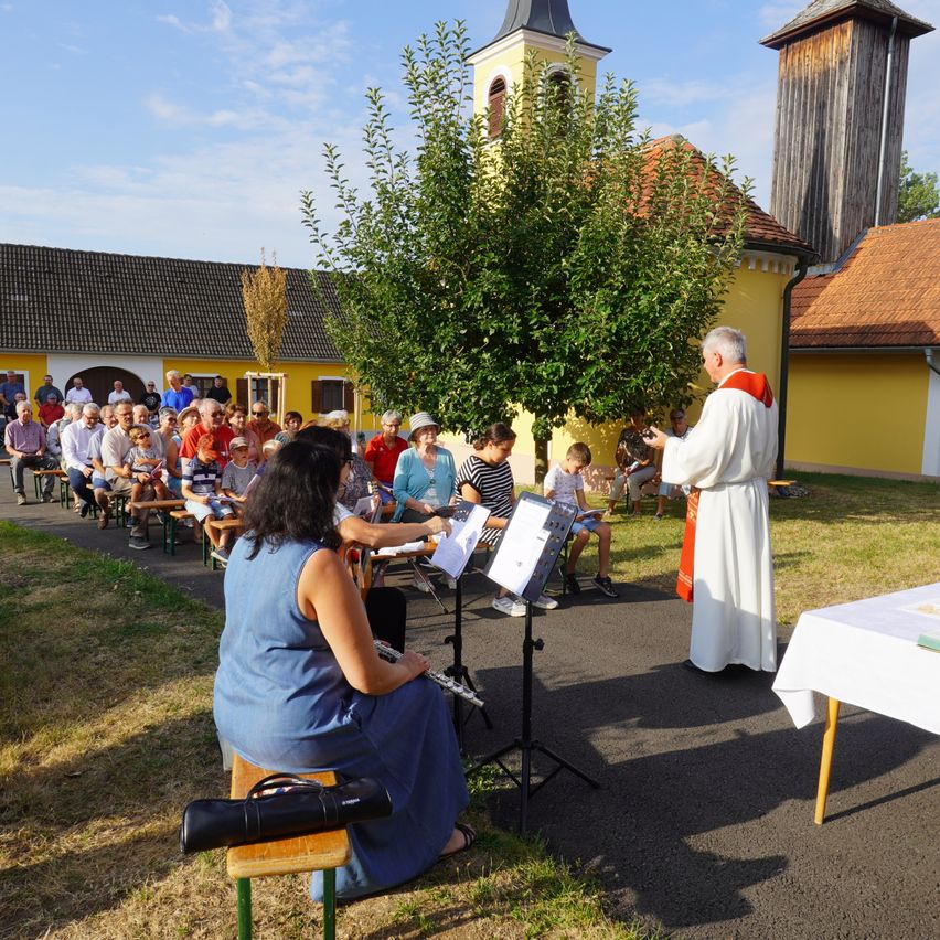 Ein Priester steht vor einer Menschenmenge, während eine Frau die Flöte spielt. Das Publikum sitzt auf Bänken und Stühlen, und ein Tisch mit einer weißen Tischdecke ist in der Nähe. Die Umgebung ist im Freien, mit einem Baum und einem gelben Gebäude im Hintergrund.