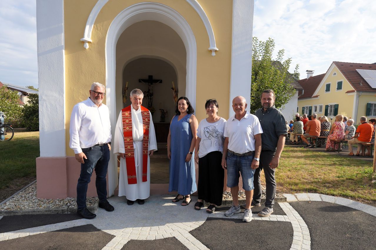 Eine Gruppe von Menschen, einschließlich eines Priesters, steht an einem sonnigen Tag vor dem Eingang einer Kirche.