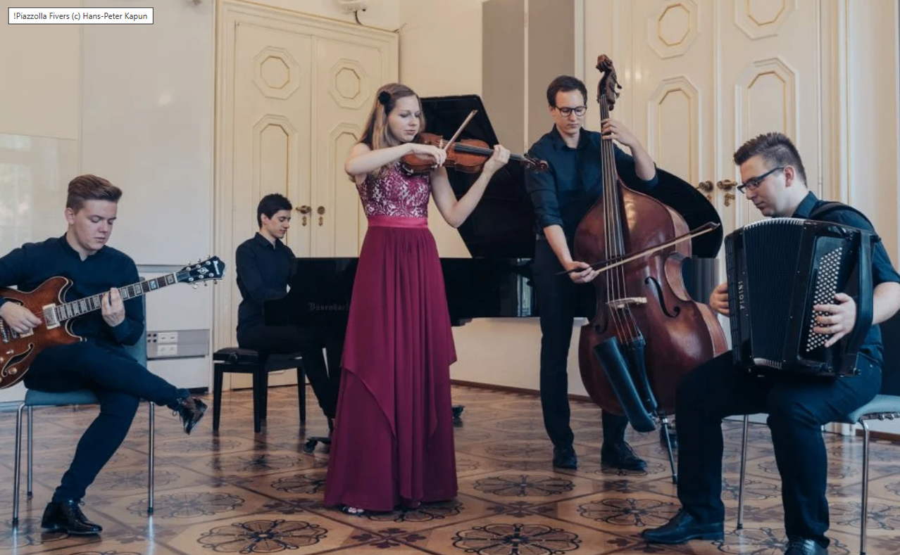 Eine Frau in einem roten Kleid spielt Geige, während ein Mann Cello spielt. Dahinter spielen zwei weitere Musiker Klavier und Gitarre.