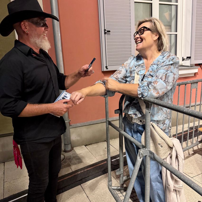 Ein Mann in einem schwarzen Cowboyhut übergibt einer Frau eine Karte, während sie lächelt und ein Telefon in der Hand hält. Sie stehen vor einer Metallbarriere.