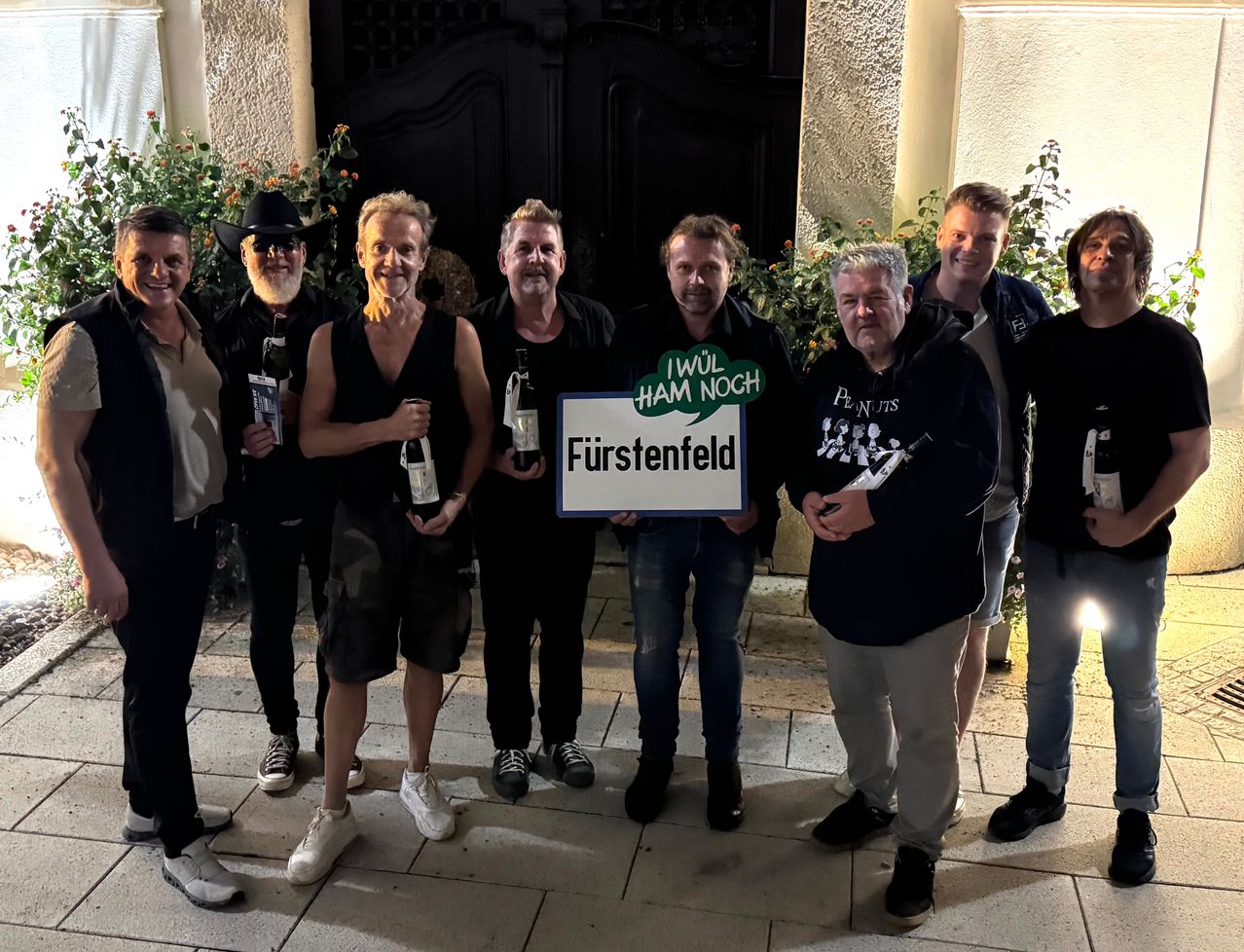 Eine Gruppe von Männern und Frauen steht draußen, lächelt und hält Weinflaschen. Ein Mann hält ein Schild mit der Aufschrift 'Fürstenfeld'.