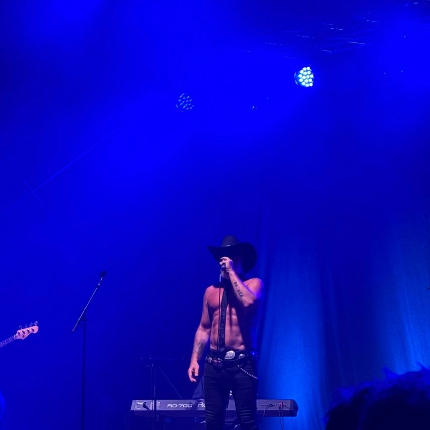 Ein oberkörperfreier Mann mit Cowboyhut tritt mit einem Keyboard auf der Bühne auf, unter blauem Licht.