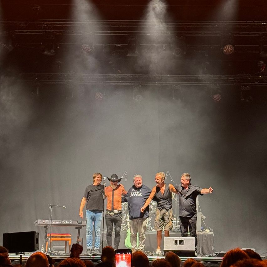 Vier Männer stehen auf einer Bühne mit Musikinstrumenten und Mikrofonen und treten vor einem Publikum unter Bühnenbeleuchtung auf.