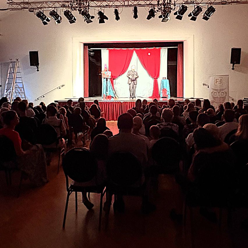 A man stands on stage in front of a red curtain, addressing an audience in a theater.