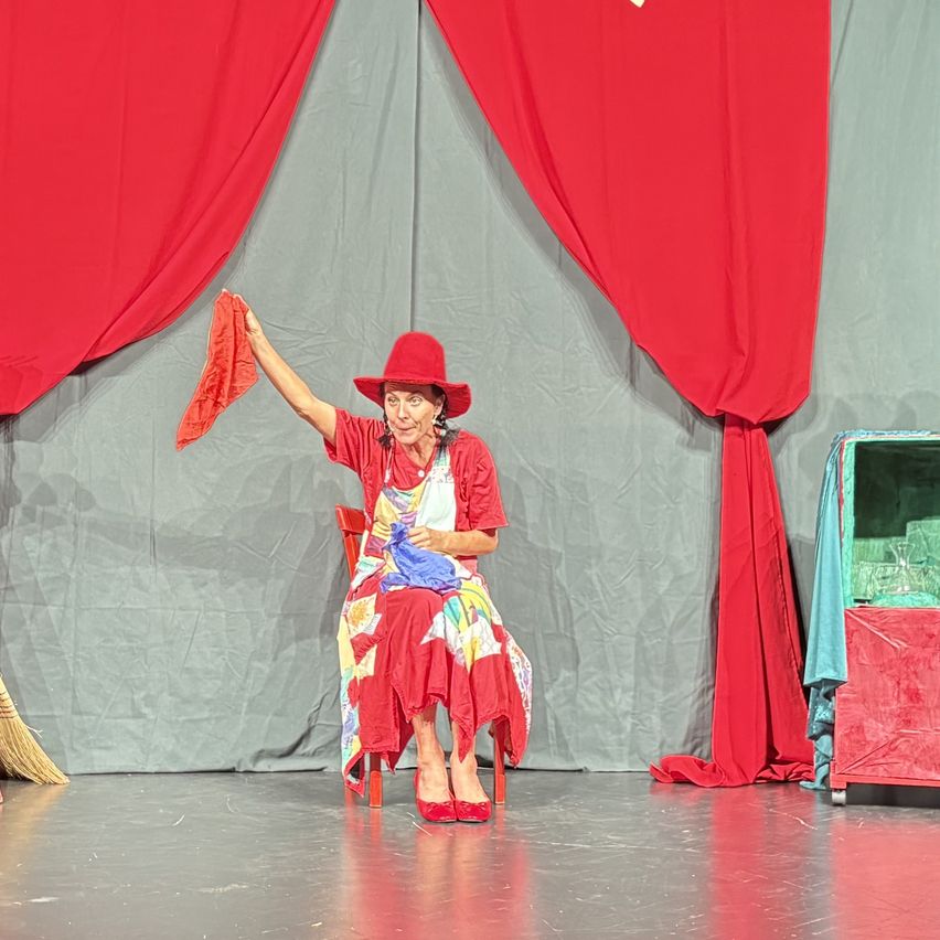 An elderly woman in a red hat and dress is sitting on a chair on a stage, waving a red cloth. Behind her, there are red curtains and a table with a blue cloth.