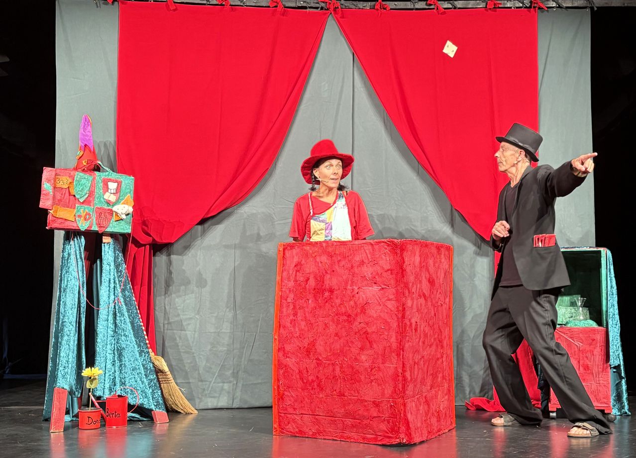 Two performers on stage, one woman in a red hat behind a red podium, and a man in a black suit and top hat. Red curtains in the background.