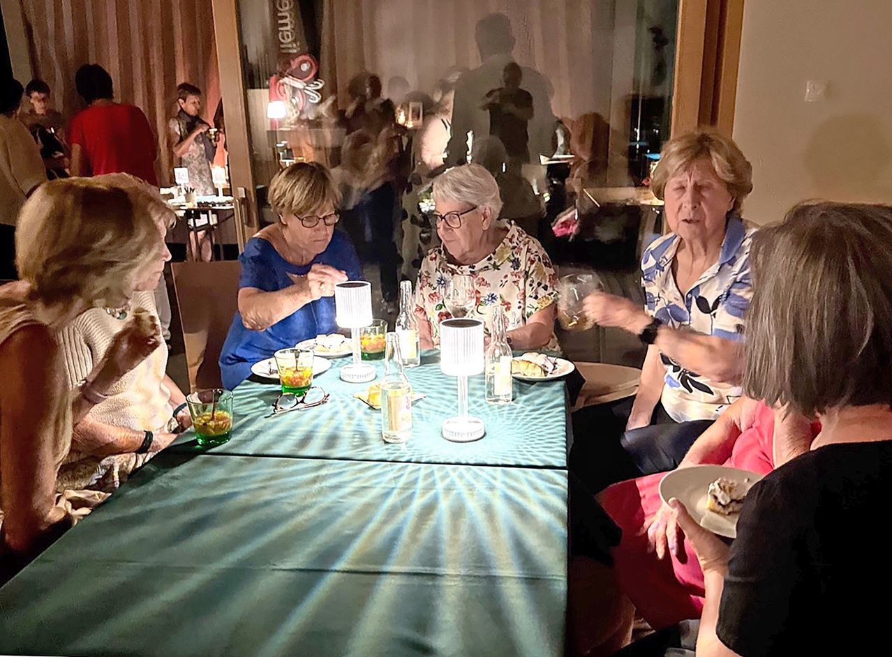 A group of older women are sitting around a table, drinking and eating. The table is set with plates, glasses, and a lamp. In the background, there are people standing and a glass wall.