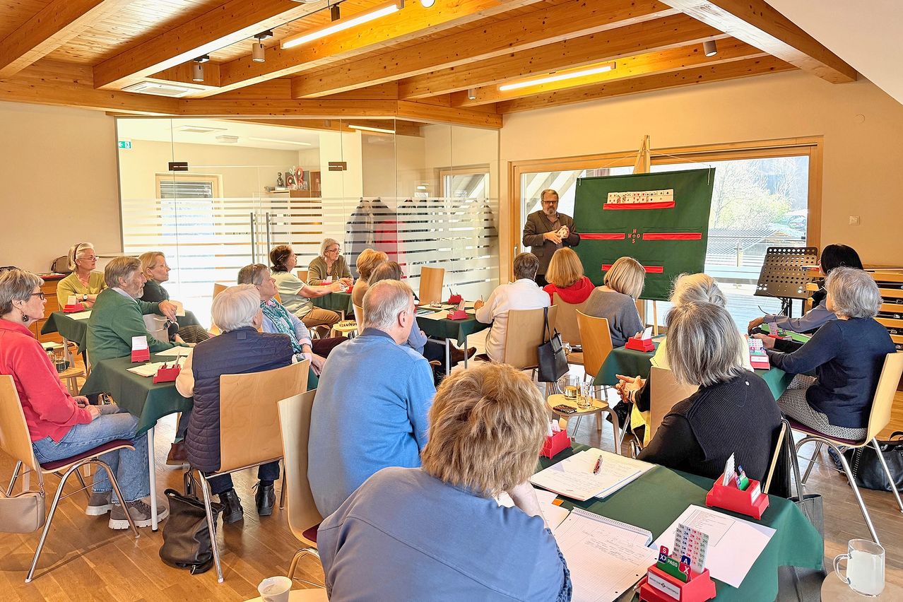 A group of people sit around a table in a room, listening to a man standing in front of a green board with a game board on it.