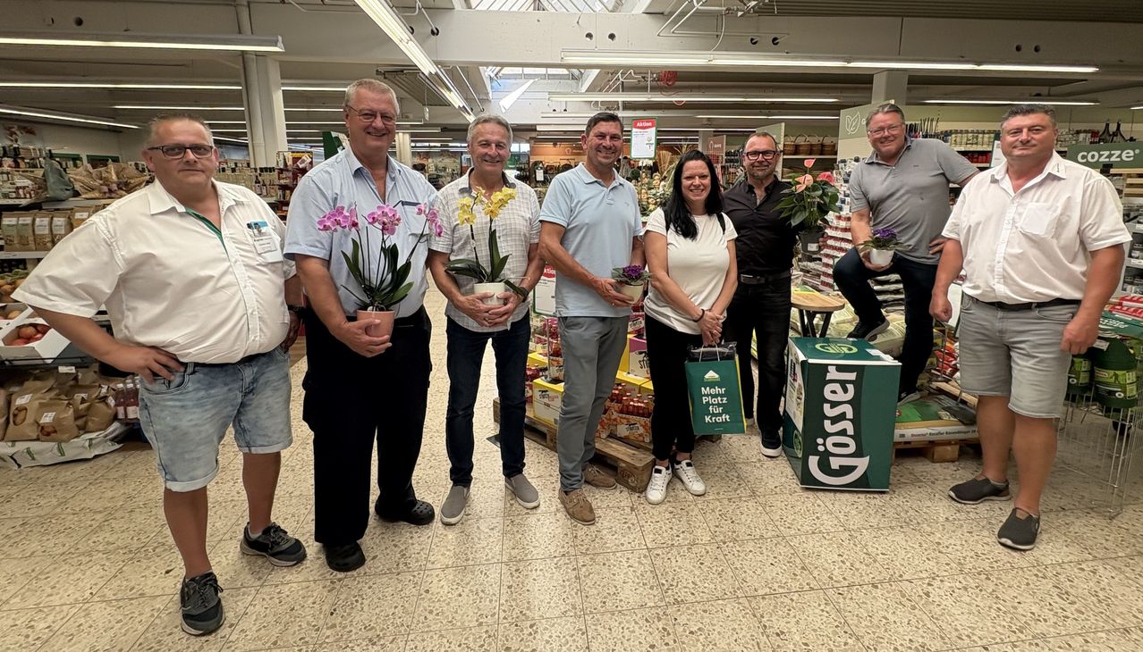A group of people in a supermarket, each holding a potted plant. They are smiling and posing for a photo. The background shows shelves with various products.