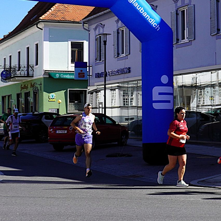 Runners in a marathon race, passing under a blue arch with the word 'Glaubandich'. Buildings and parked cars are in the background.