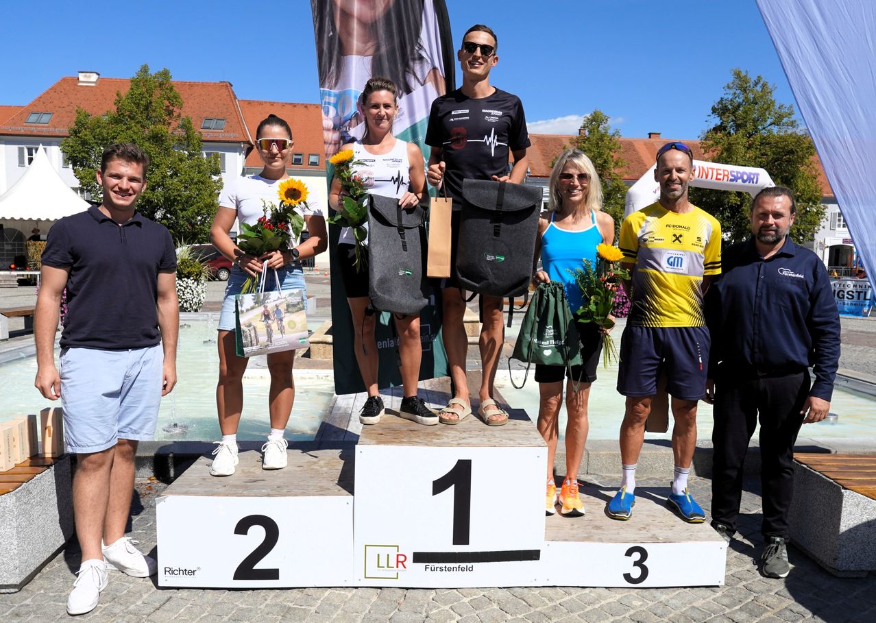 A group of people stand on a podium with flowers and bags, likely celebrating a cycling event. The podium has numbers 1, 2, and 3. A man in the middle holds a black bag.
