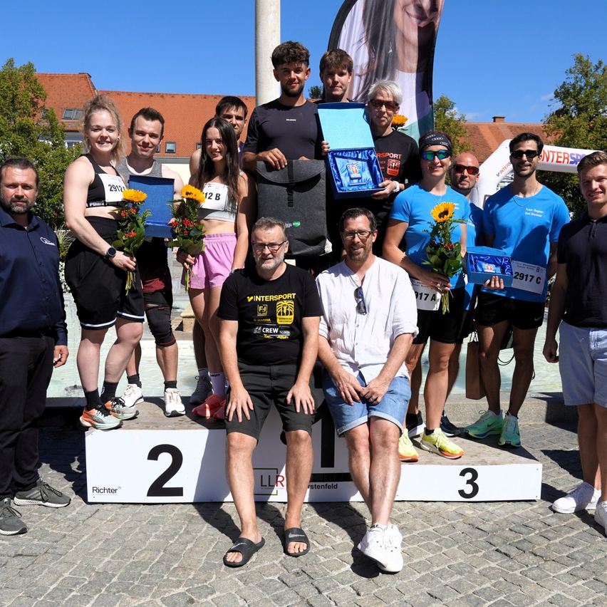 A group of people stand on a podium, holding flowers and awards. They are dressed in athletic wear and appear to be celebrating a victory. The podium has numbers 2 and 3 on it.