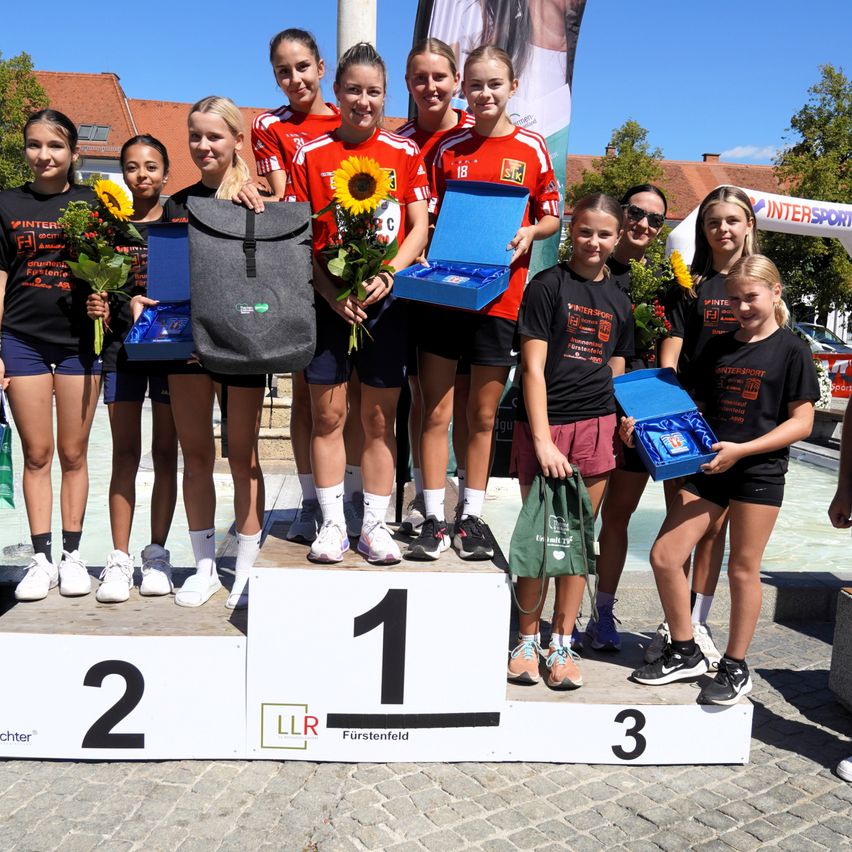 A group of young women stand on a podium with flowers and awards, wearing matching red and black uniforms. The podium has numbers 1, 2, and 3 on it.